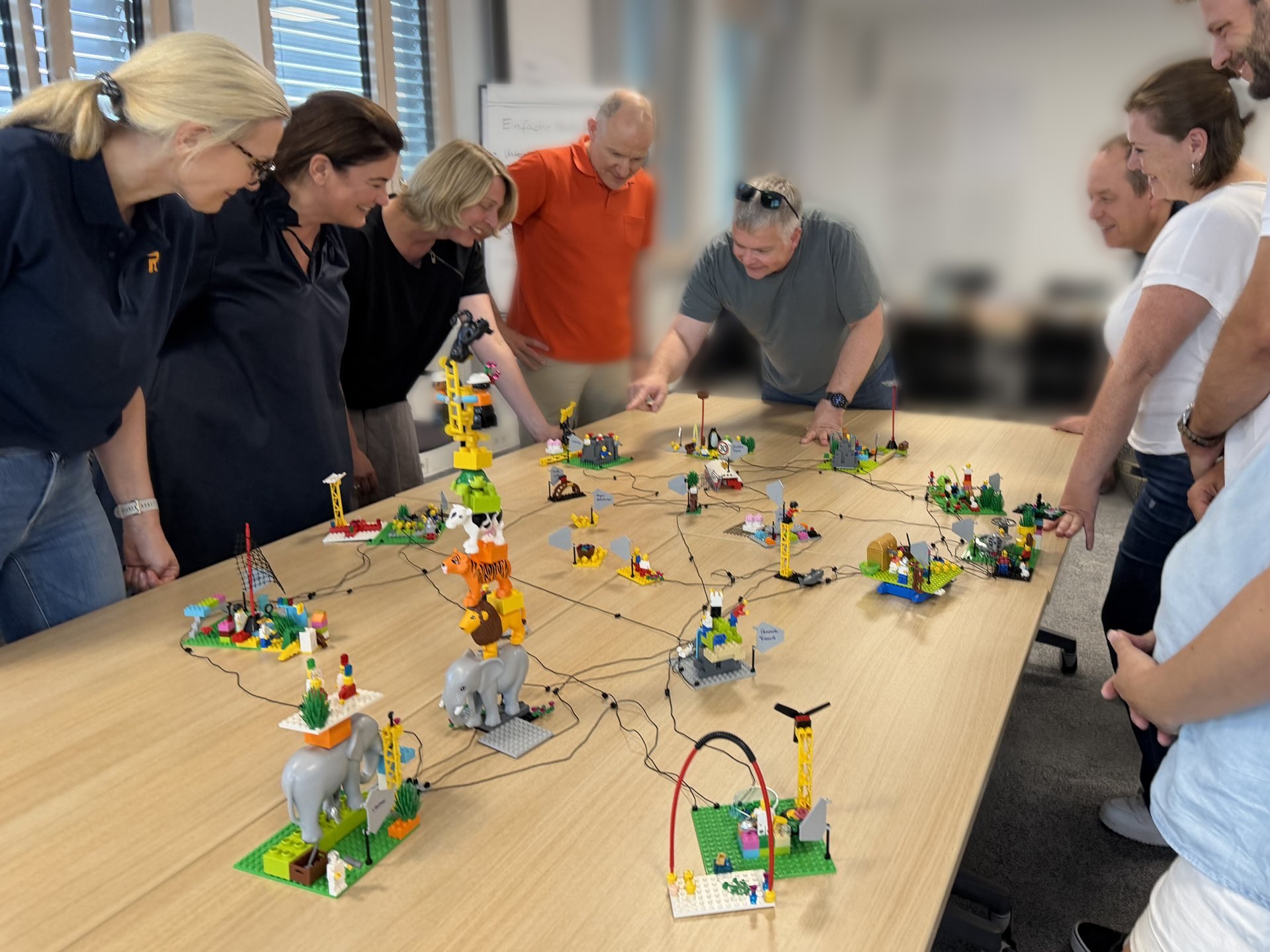 Zehn Teilnehmende stehen um einen langen Tisch mit einer Landschaft aus LEGO-Modellen, die mit Schnüren verbunden sind. In der Mitte zeigt ein Mann im grauen T-Shirt auf ein Modell.