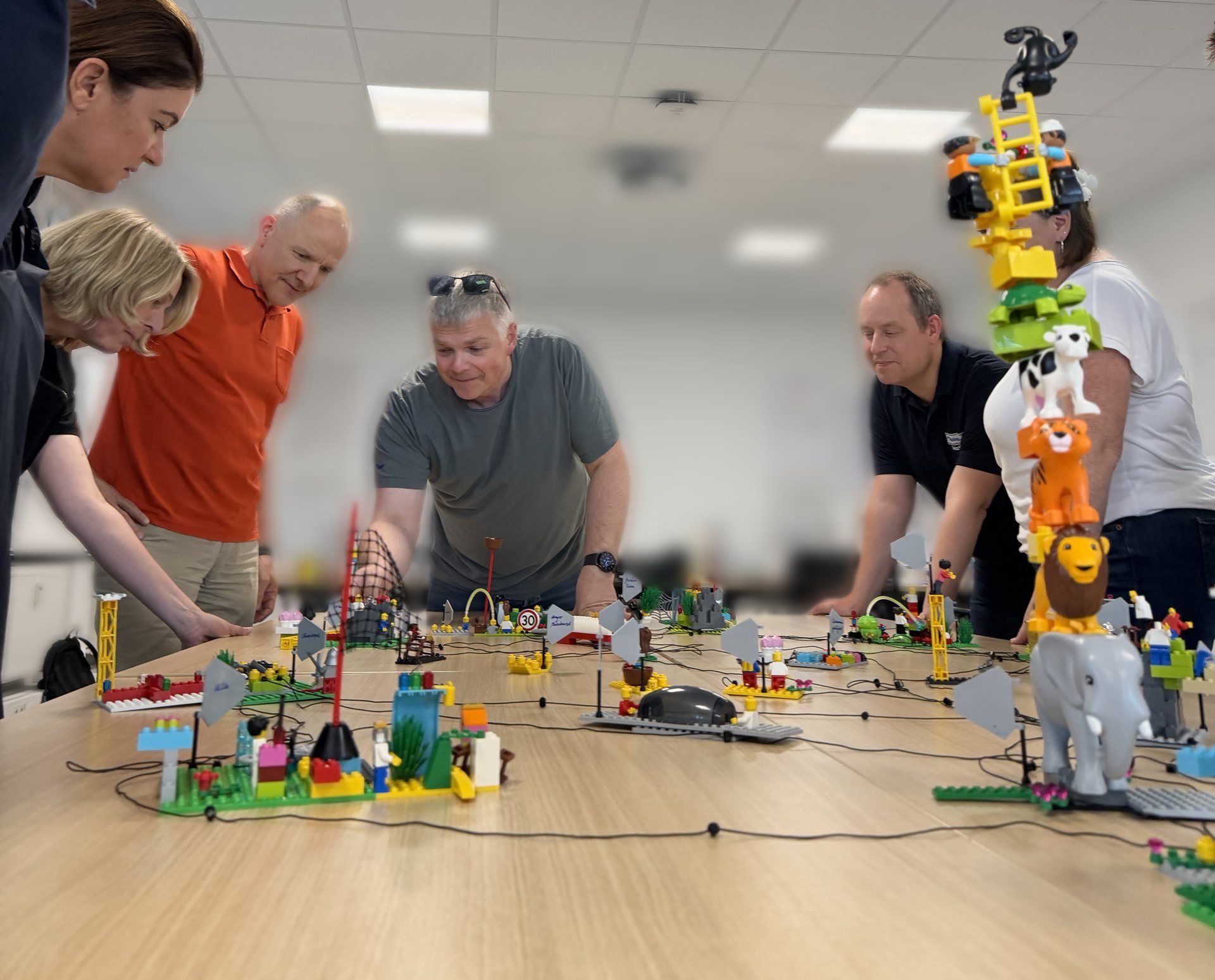Blick über einen Tisch mit verbundenen LEGO-Modellen, vorne ein Turm aus Tierfiguren, dahinter die Gruppe, die sich über die Modelle beugt.