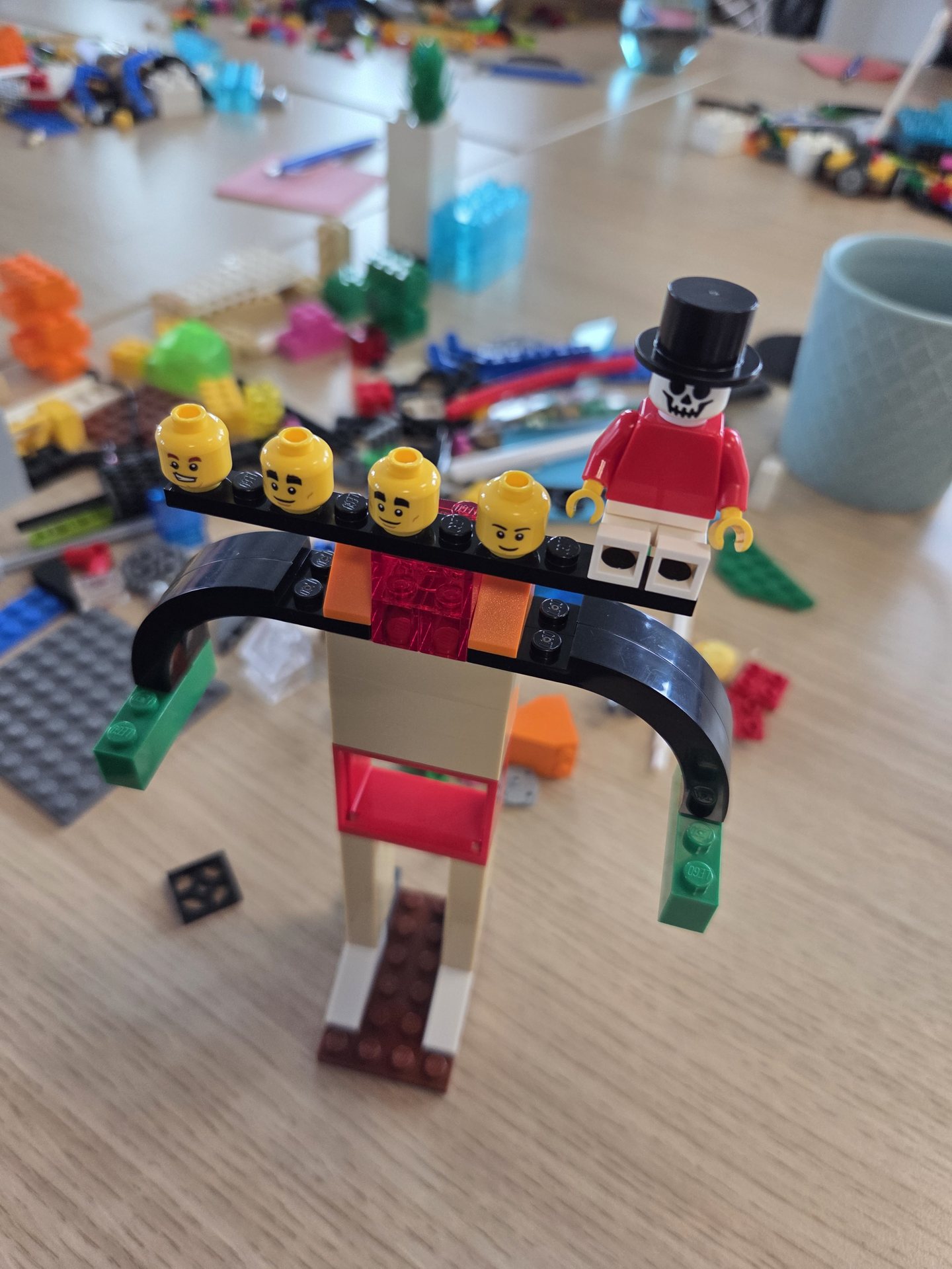 LEGO-Modell: ein Bogen trägt einen Balken mit vier Minifigurköpfen, daneben sitzt eine Figur mit Zylinder